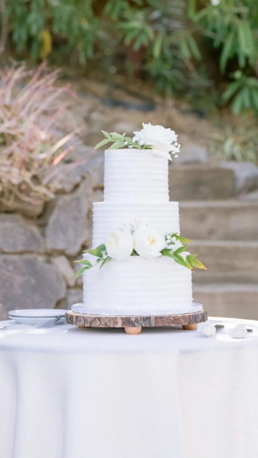 Three-tier white textured cake with white peonies and greenery on a wood slice stand outdoors