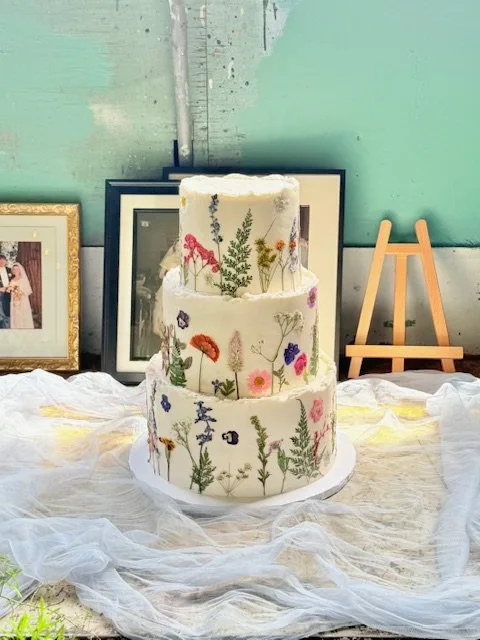 Three-tier white cake covered in pressed edible wildflowers and ferns against a mint green wall